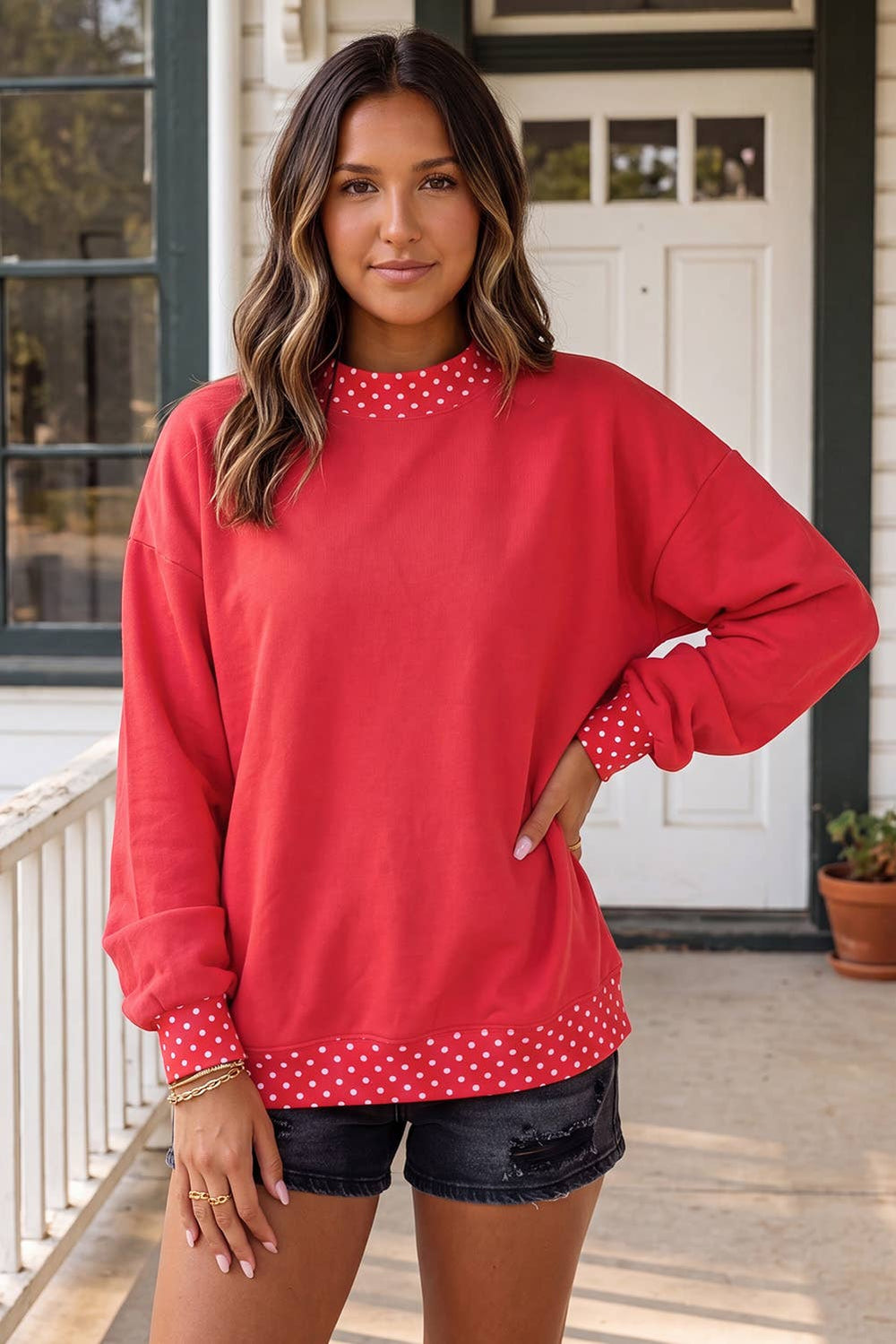 A woman wearing a red long-sleeve sweatshirt with white polka dot trim at the mock neck and hem, paired with black denim shorts, standing on a front porch.