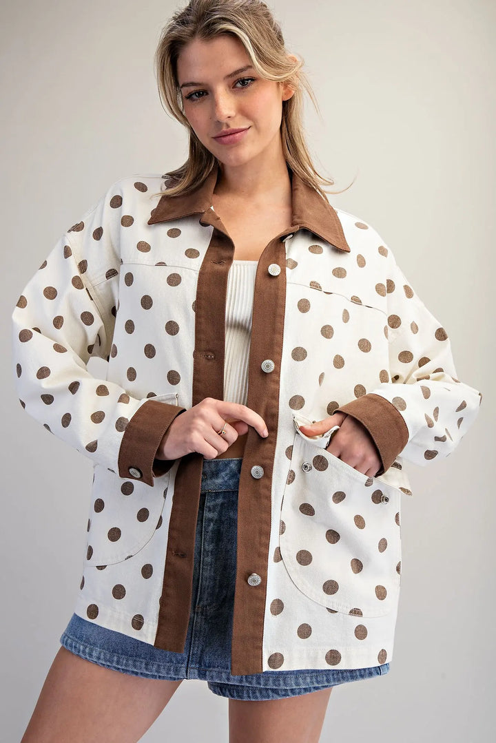 A woman wearing a white button-up shirt jacket with brown polka dots and solid brown collar, placket, and cuffs, styled with denim shorts over a white ribbed top against a light gray studio background.