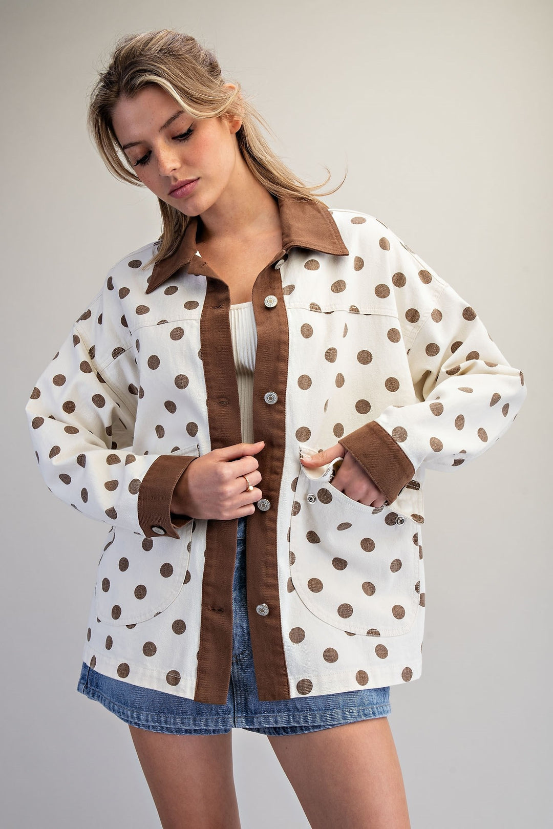 A woman wearing a white button-up shirt jacket with brown polka dots and solid brown trim, styled with a white top and denim shorts against a neutral studio background.