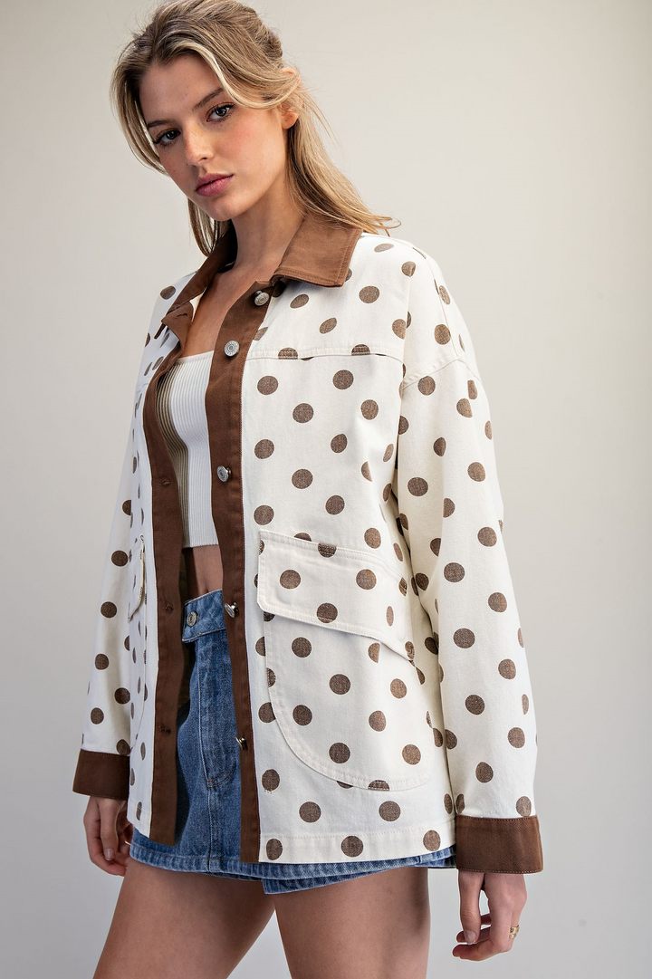 A woman models a white button-up jacket with brown polka dots and solid brown trim, worn open over a white crop top and blue denim shorts against a plain beige studio background.