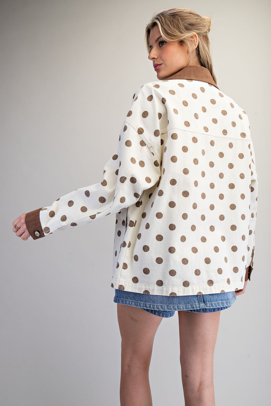 A woman shown from the back wearing a cream-colored jacket with brown polka dots and a brown collar, paired with denim shorts, against a neutral gray background.