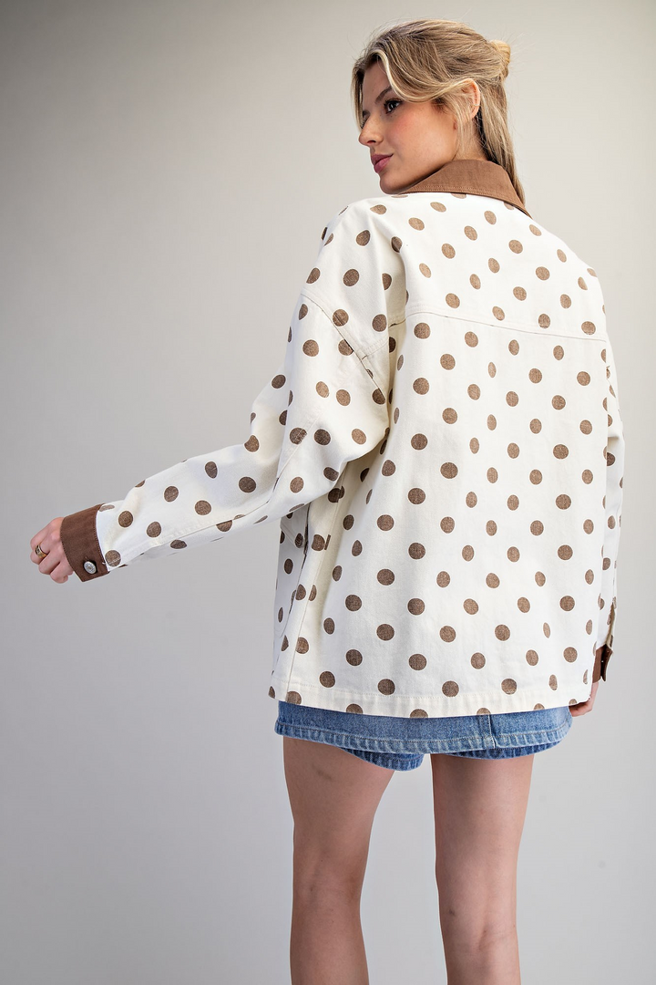 A woman shown from the back wearing a cream-colored jacket with brown polka dots and a brown collar, paired with denim shorts, against a neutral gray background.