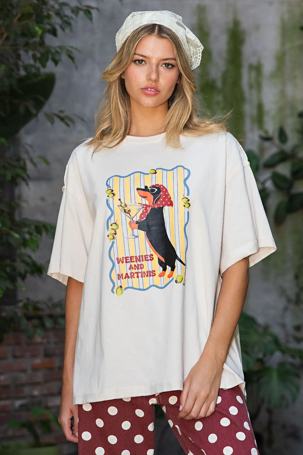 A woman wearing an oversized cream graphic t-shirt with a dachshund illustration and the words 'WEEZIES AND MARTINS', paired with red and white polka dot pants and a white lace headband, set against a blurred outdoor background.
