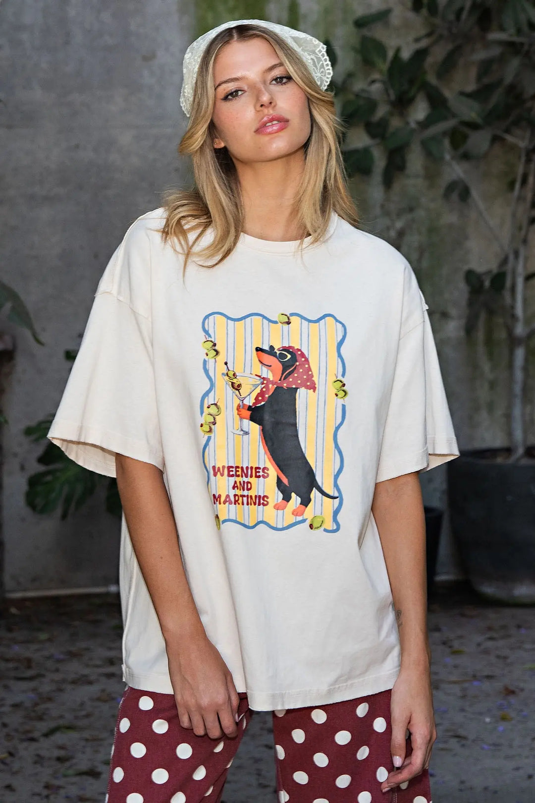 A woman wearing an oversized cream-colored graphic t-shirt with a dachshund print, paired with red and white polka dot pants and a white lace headband, standing in an outdoor patio setting.
