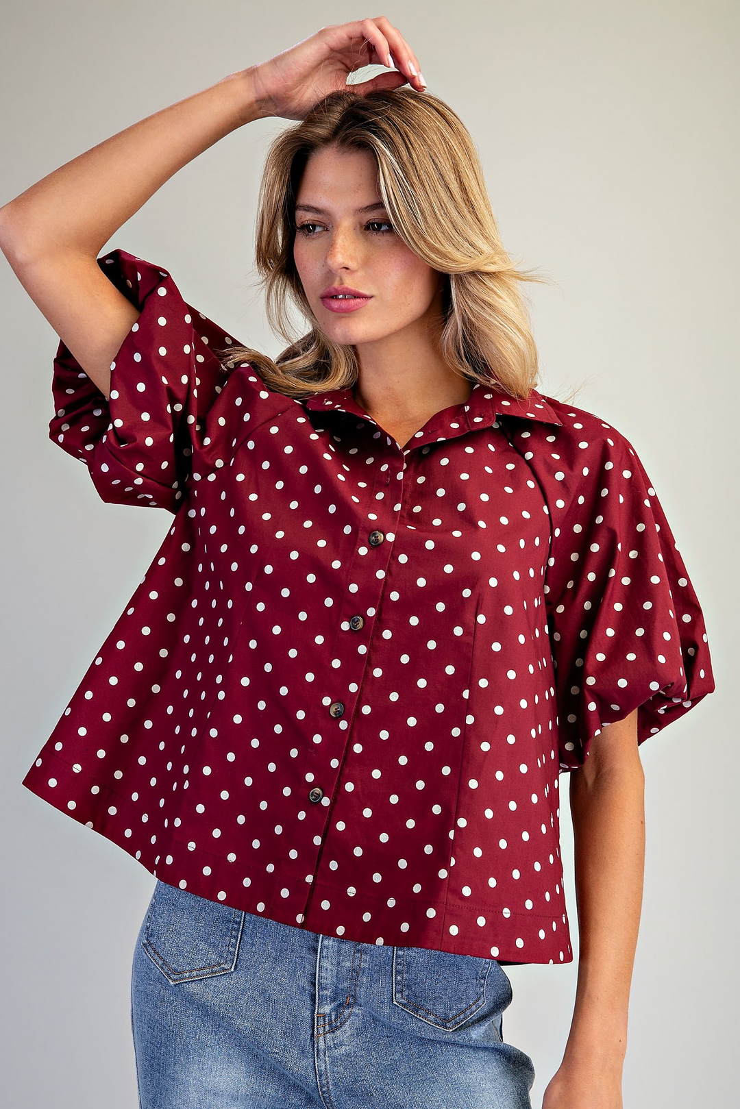 A woman wearing a burgundy short-sleeve button-up blouse with white polka dots and light blue jeans, posing against a neutral light gray background.