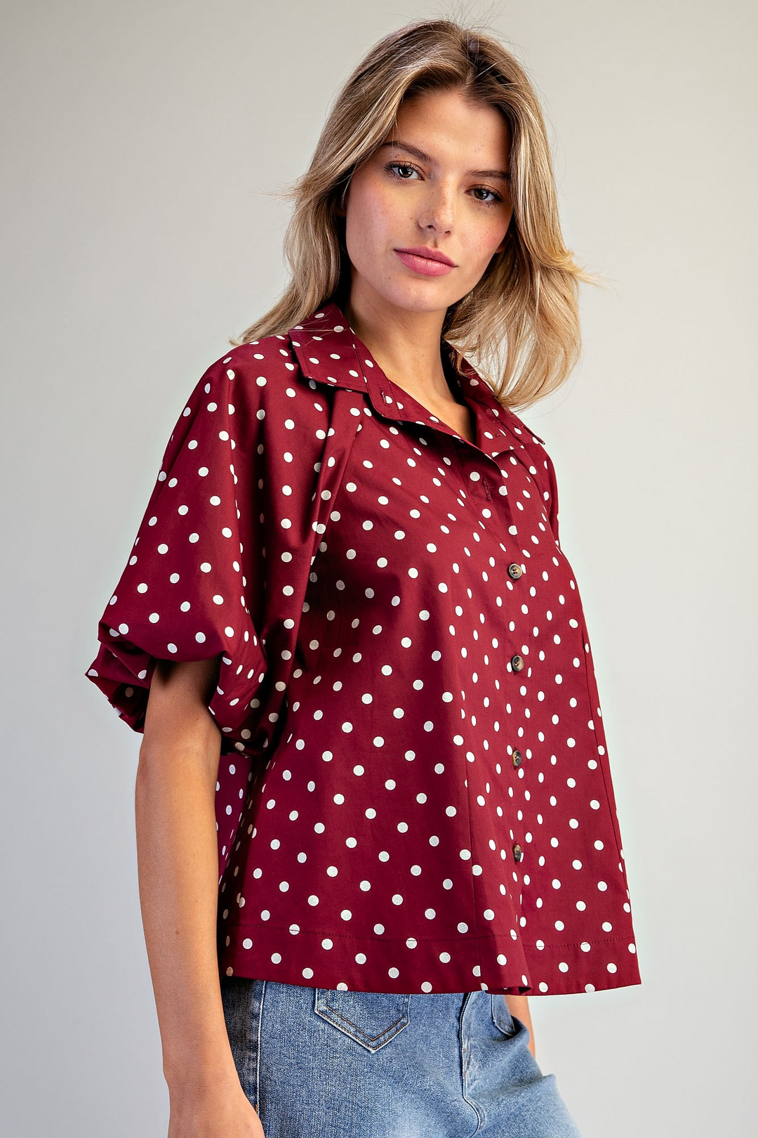 A woman wearing a burgundy short-sleeve button-up blouse with white polka dots and light blue jeans, posing against a neutral light gray background.