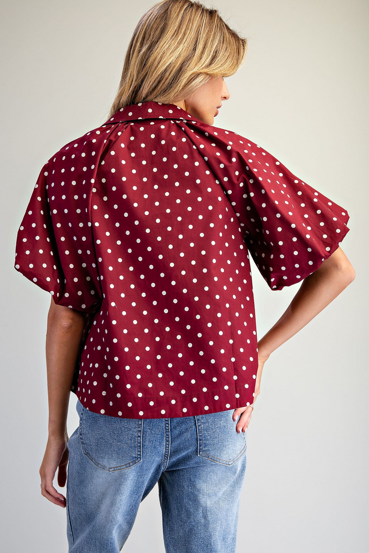 A woman shown from the back wearing a short-sleeve collared blouse with white polka dots on a deep red base, paired with light blue jeans, against a plain light gray studio background.