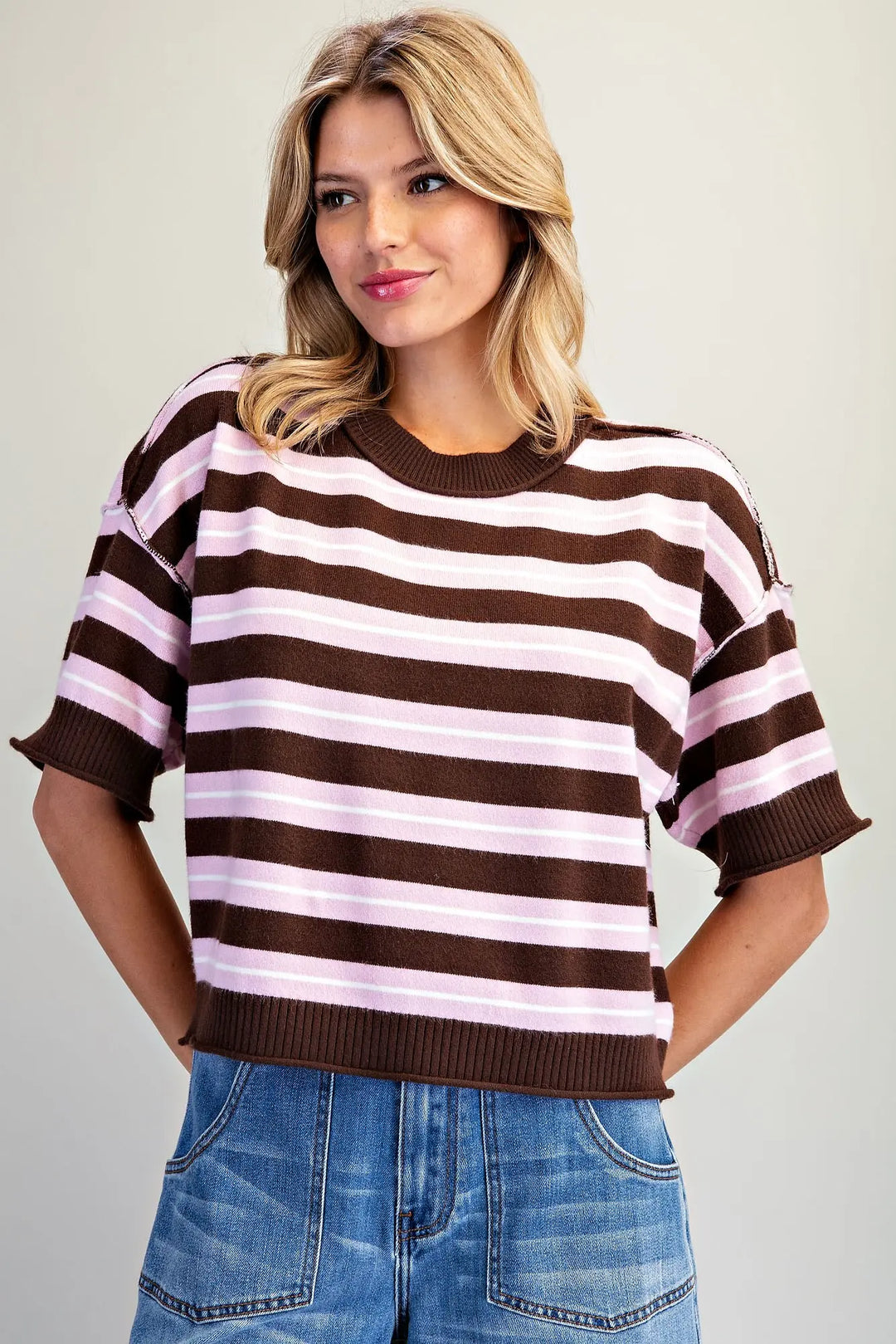 A woman wearing a brown and light pink horizontally striped short-sleeve knit top with blue jeans, photographed against a plain light gray studio background.