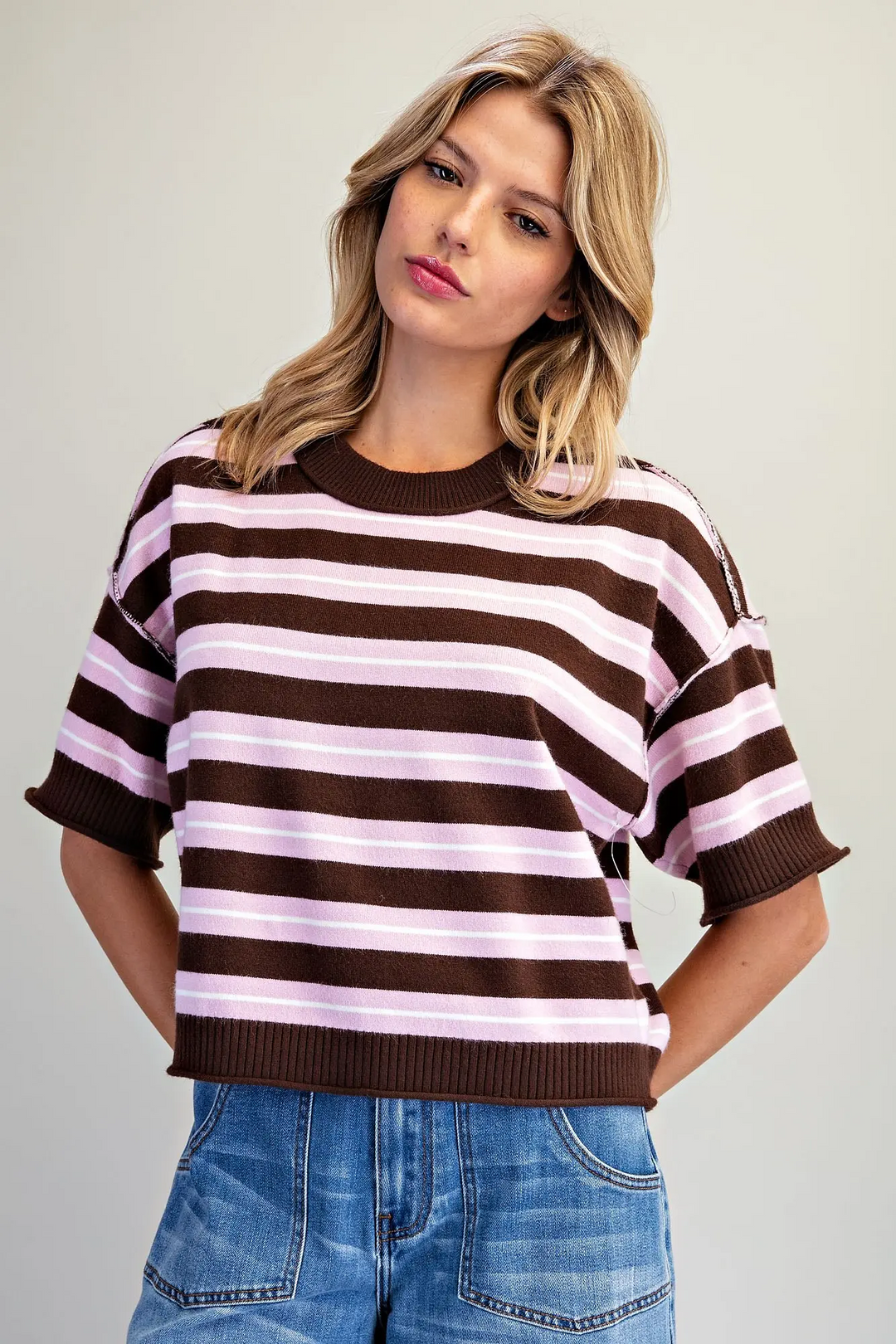 A woman wearing a brown and light pink horizontally striped short-sleeve knit top with ribbed collar, cuffs, and hem, paired with blue jeans, against a plain light gray studio background.