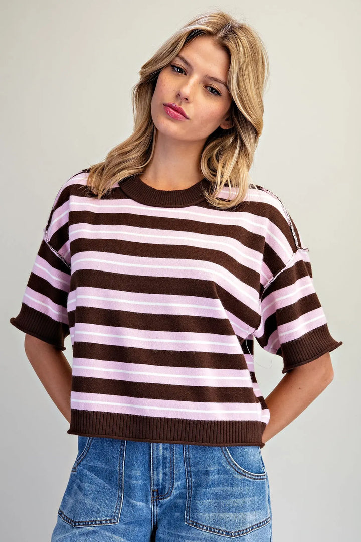 A woman wearing a brown and light pink horizontally striped short-sleeve knit top with ribbed collar, cuffs, and hem, paired with blue jeans, against a plain light gray studio background.