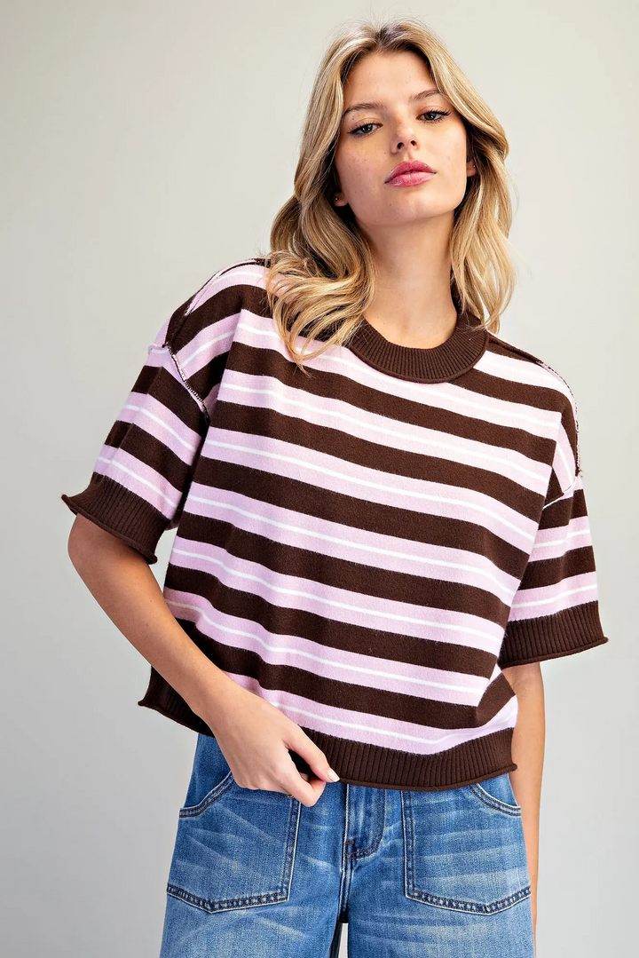A woman wearing a brown and light pink horizontally striped short-sleeve knit top with ribbed cuffs and collar, paired with blue jeans, against a plain light gray background.