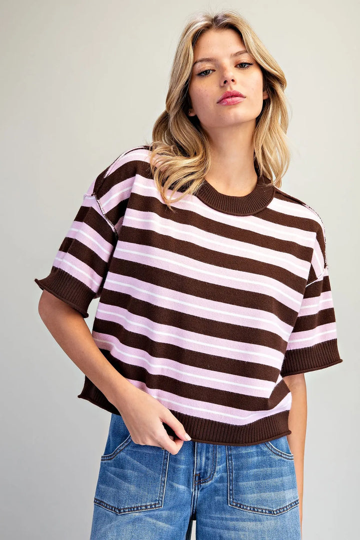 A woman wearing a brown and light pink horizontally striped short-sleeve knit top with ribbed cuffs and collar, paired with blue jeans, against a plain light gray background.