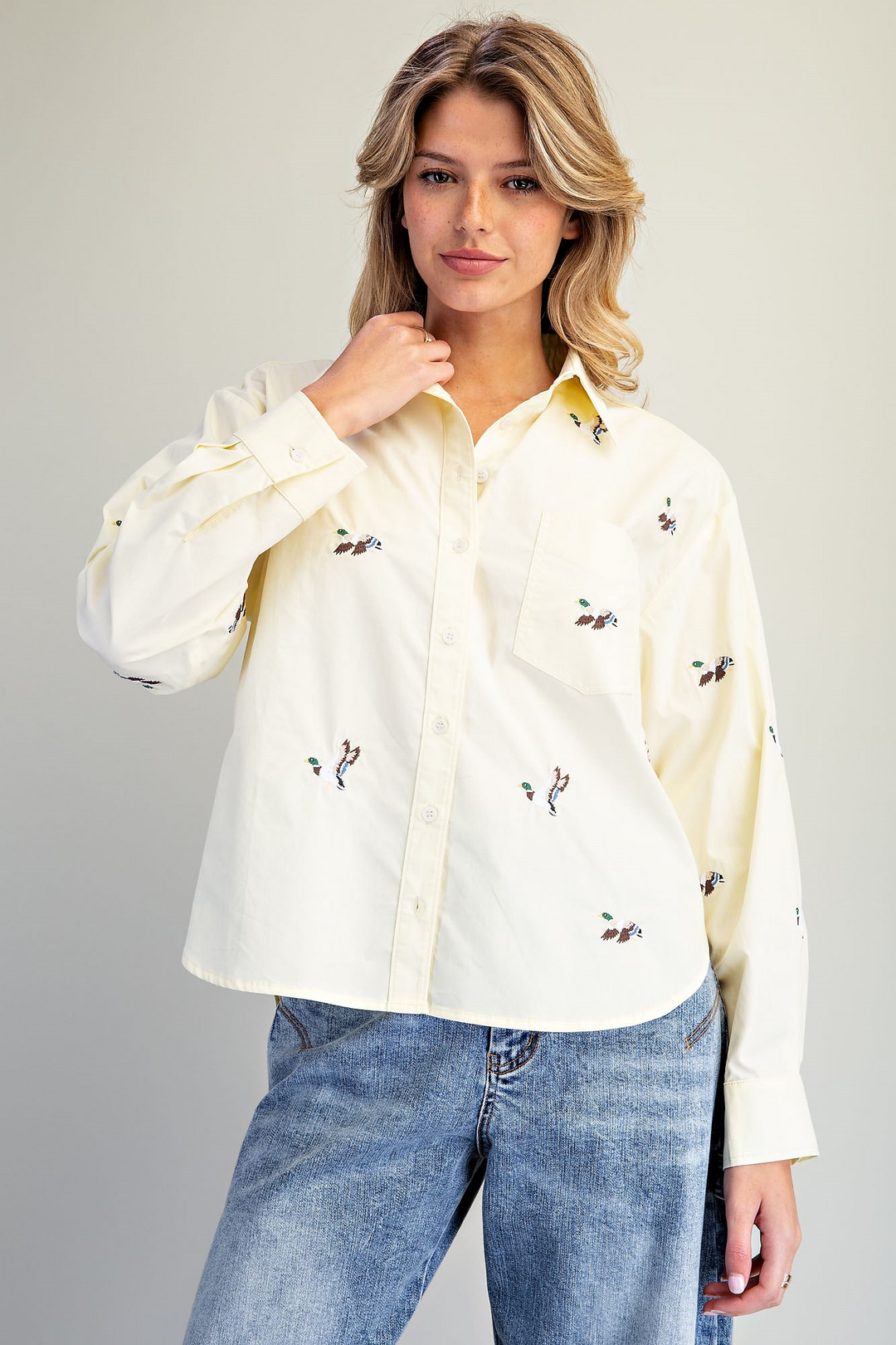 A woman wearing a cream-colored long-sleeve button-up shirt with small embroidered motifs, paired with blue jeans, against a neutral light gray background.