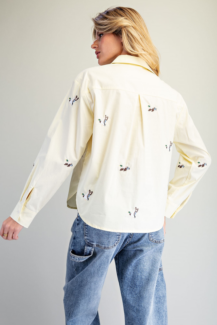 A woman shown from the back wearing a cream-colored long-sleeve button-up shirt with small black embroidered details, paired with blue jeans, against a plain light gray background.
