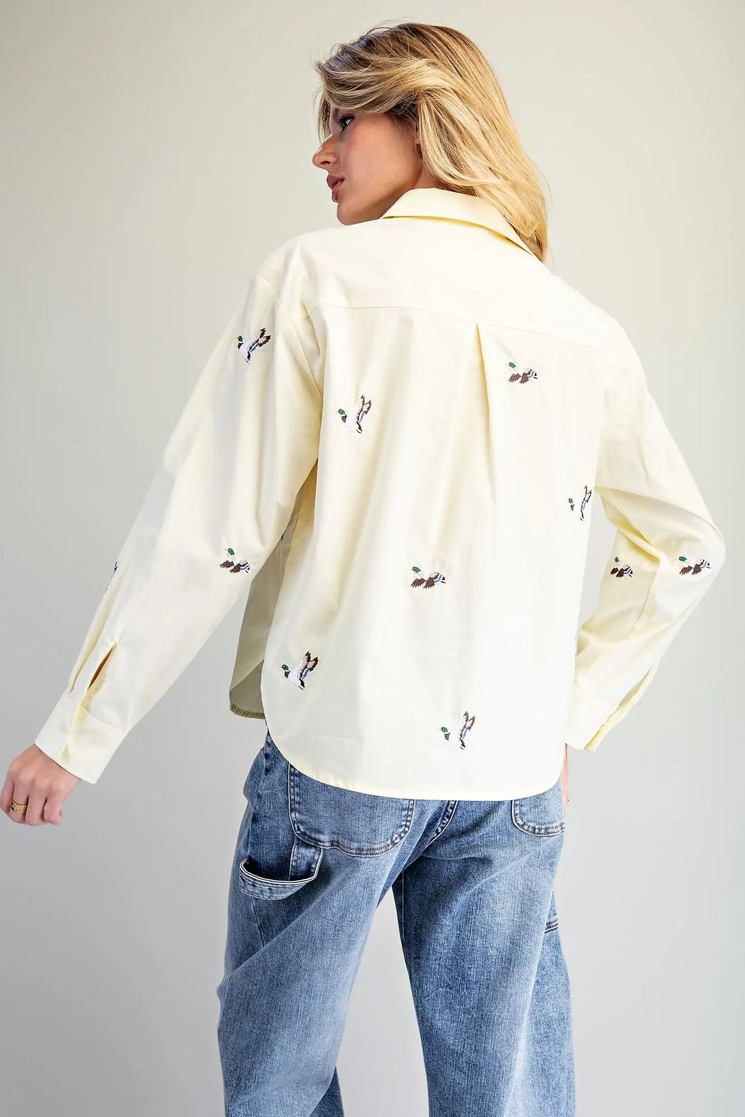 A woman shown from the back wearing a cream-colored long-sleeve button-up shirt with small black embroidered details, paired with blue jeans, against a plain light gray studio background.