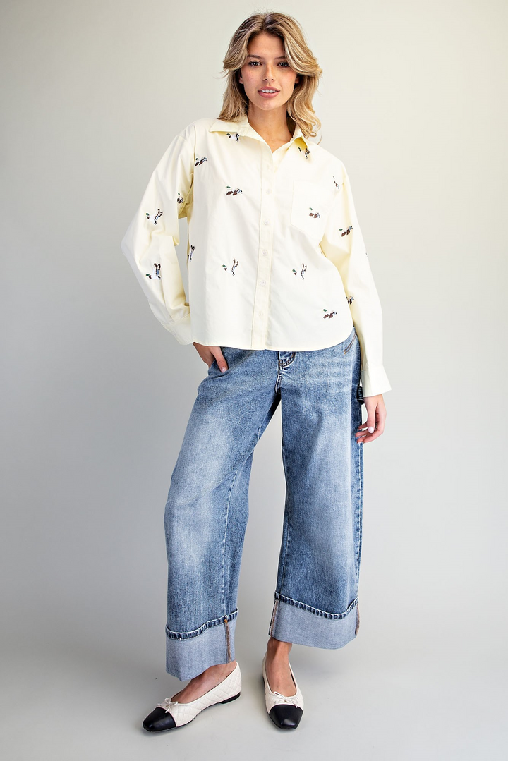 A woman modeling a cream-colored long-sleeve button-up shirt with small black embroidered motifs, paired with wide-leg light-wash blue jeans and black-and-white pointed flats, against a neutral gray studio background.