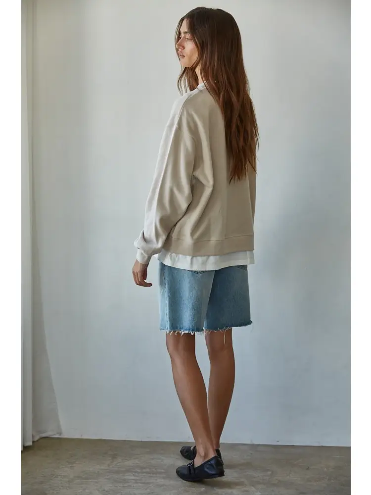A woman stands in profile wearing an oversized beige sweatshirt layered over a white collared shirt, paired with a light blue denim skirt and dark loafers, set against a plain light gray wall.