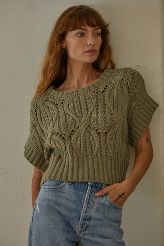 Woman wearing a green knitted sweater and blue jeans against a plain wall.
