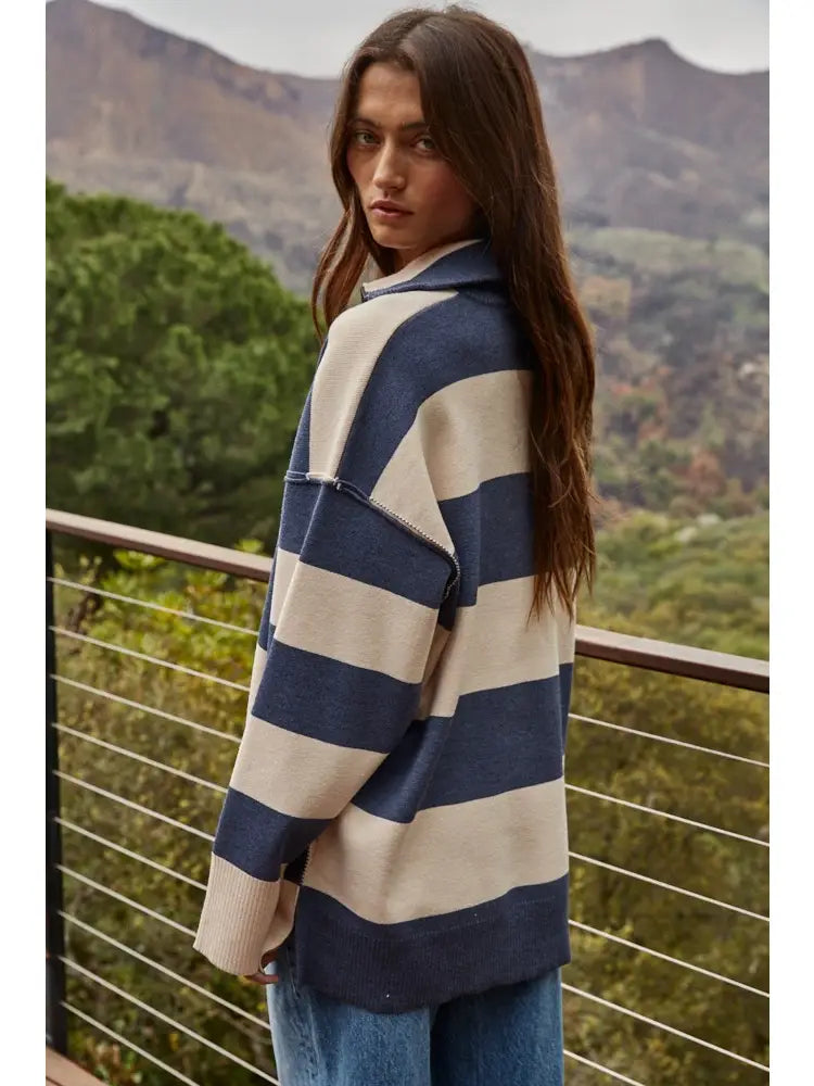 A woman wearing a blue and cream striped knit cardigan with a hood, standing on a balcony with a mountain and forest background.