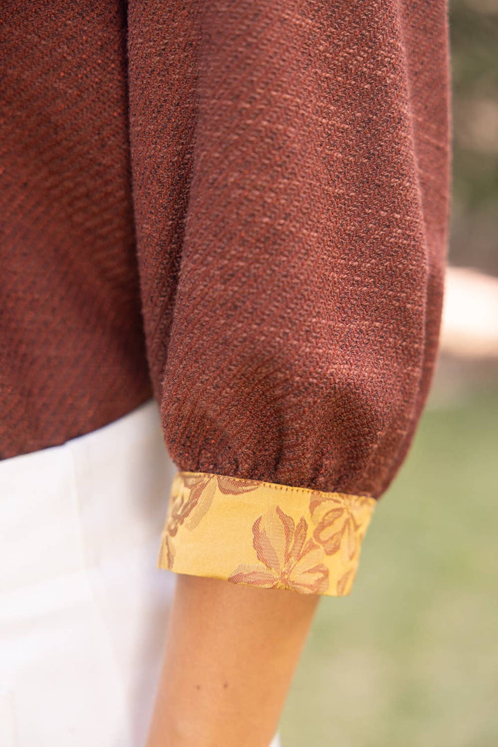 Close-up of a person wearing a rust-brown textured long-sleeve top with a yellow patterned cuff, paired with white bottoms, against a blurred outdoor background.