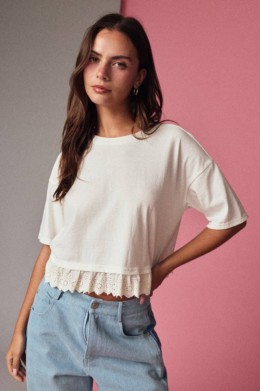 Woman wearing a white top with lace details and light blue jeans against a pink and gray background