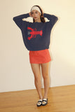 Person wearing a navy sweater with a red lobster design and a red skirt on a plain background