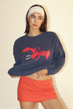 Woman wearing a navy sweater with a red lobster design and red striped shorts on a beige background