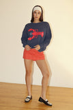 Woman wearing a navy sweater with a red lobster design and red striped shorts on a plain background