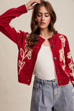 Woman wearing a red cardigan with white patterns, white top, and blue jeans on a beige background