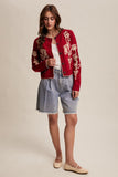 Woman wearing a red embroidered cardigan with denim shorts on a beige background