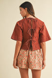 Woman wearing a rust-colored top with lace-up back and floral shorts on a beige background