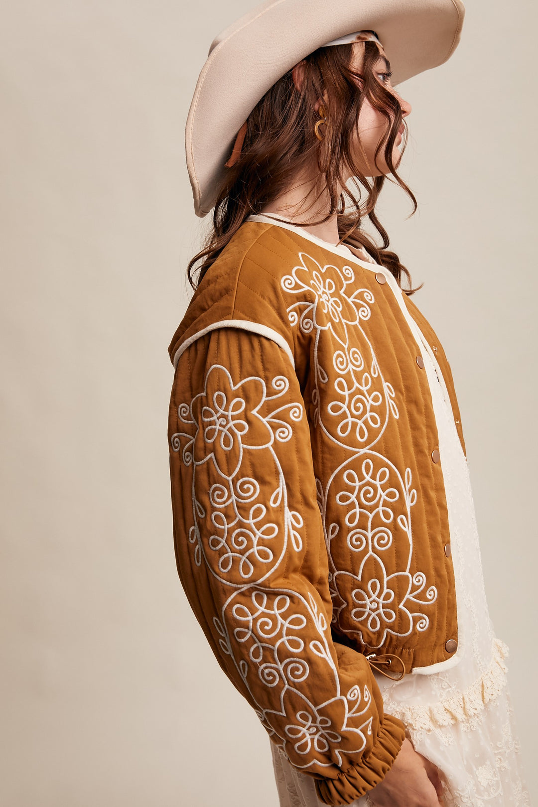Woman wearing a brown embroidered jacket and white hat against a beige background