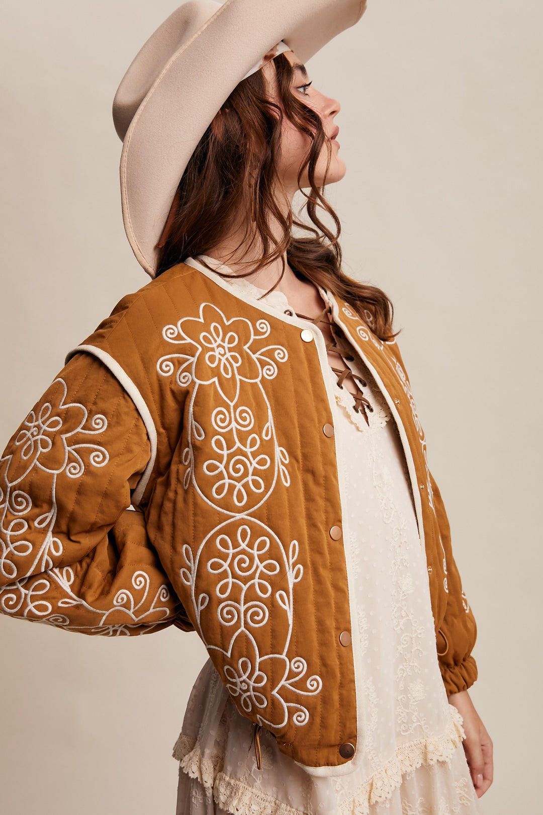 Woman wearing a brown embroidered jacket and beige hat against a beige background