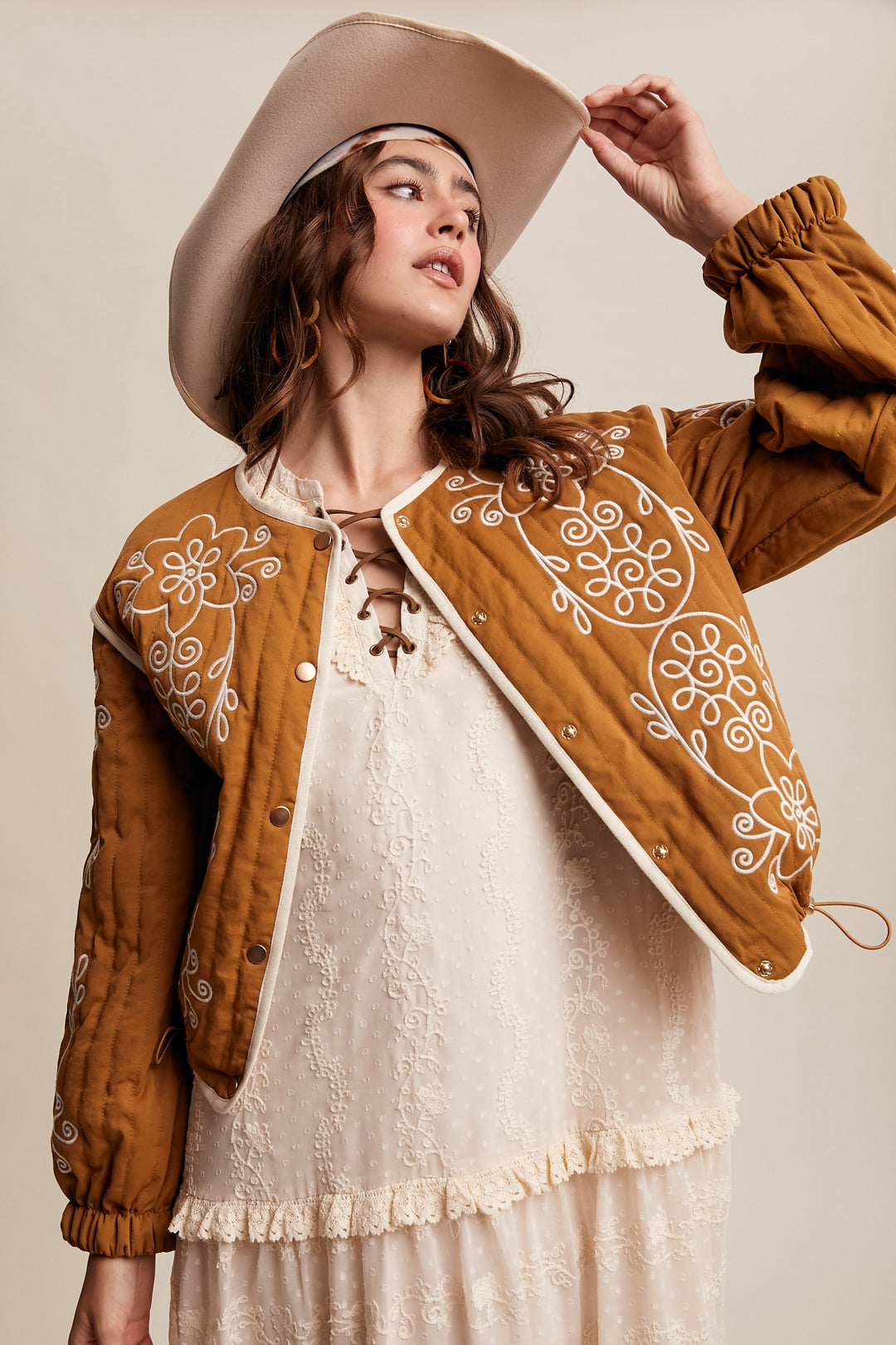 Woman wearing a brown embroidered jacket and beige hat against a neutral background