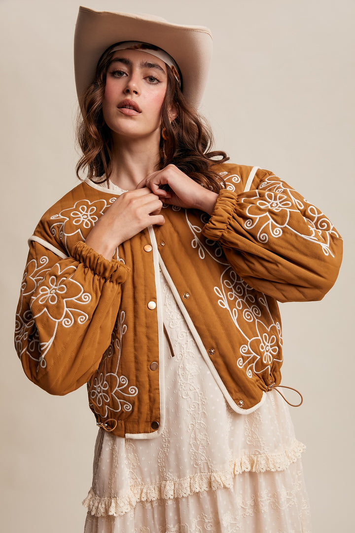 Woman wearing a brown embroidered jacket and beige hat against a neutral background