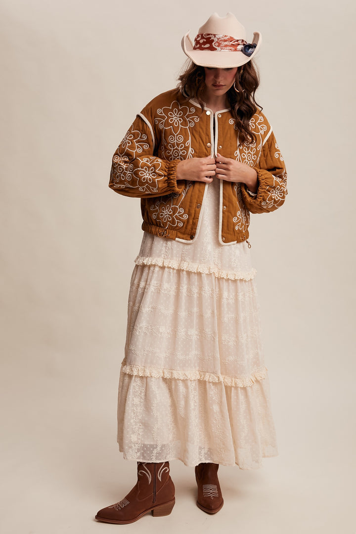 Person wearing a brown embroidered jacket, white dress, and cowboy hat on a beige background