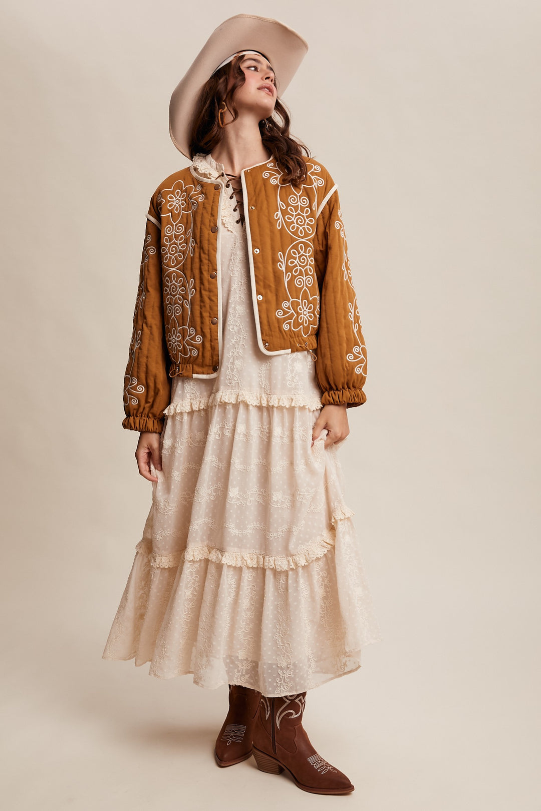 Woman wearing a brown embroidered jacket, white dress, and brown boots on a beige background