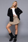Woman wearing a black jacket, white shirt, beige skirt, and black boots on a gray background