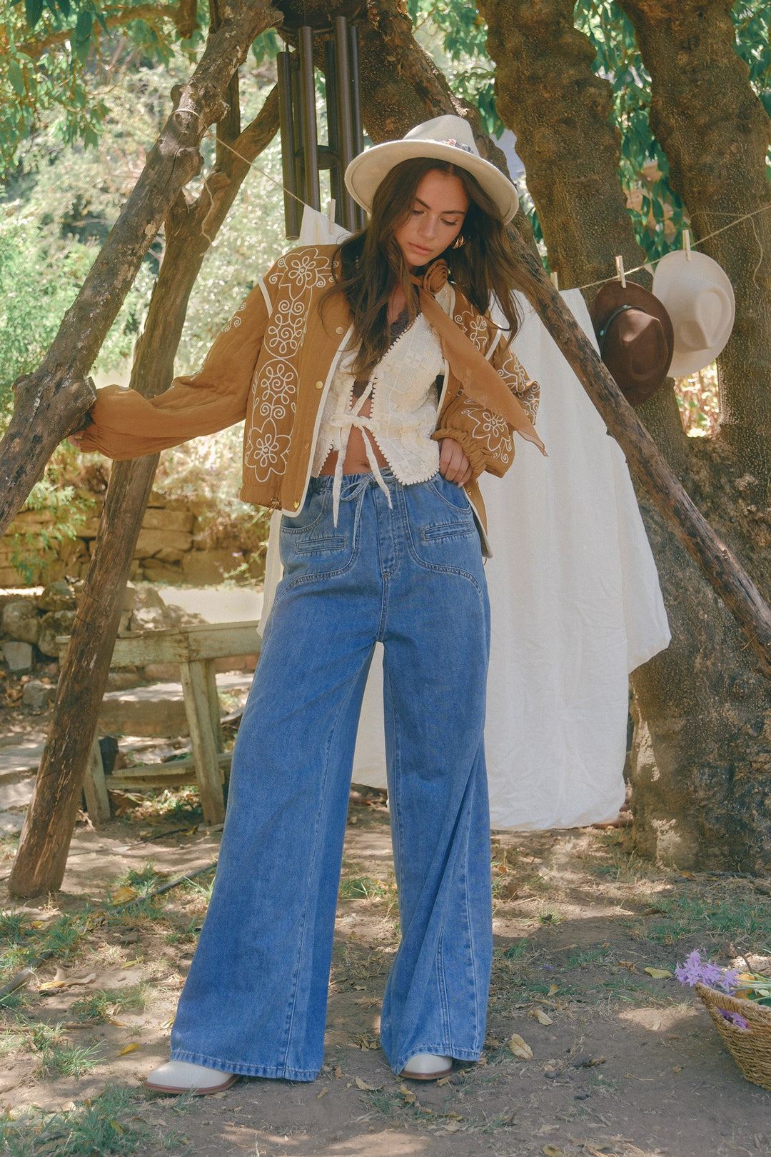 Woman in a rustic outdoor setting wearing a brown embroidered jacket, white top, blue jeans, and a white hat.