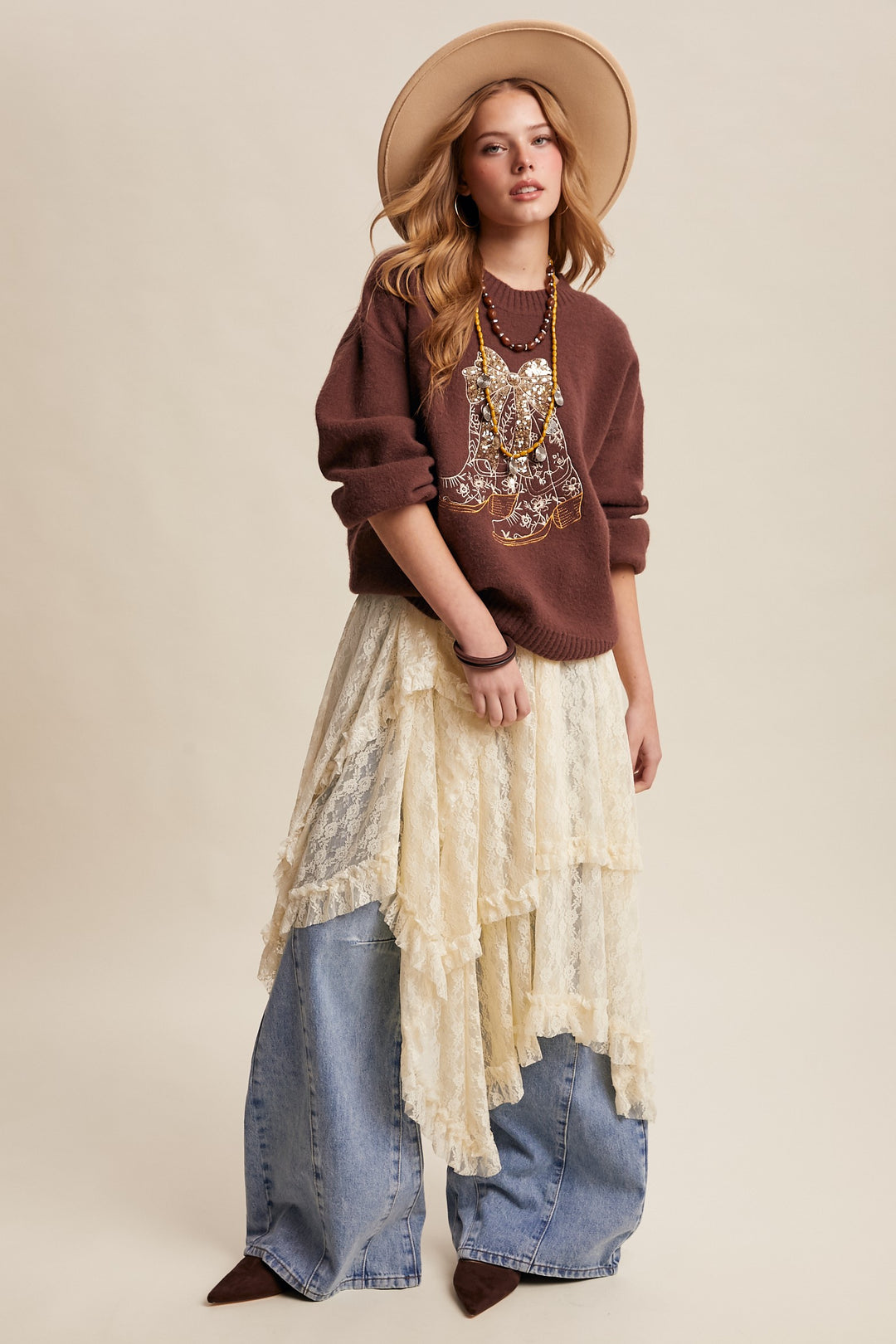 Woman wearing a brown sweater with intricate design, beige lace top, blue jeans, and a wide-brimmed hat on a beige background.