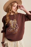 Woman wearing a brown sweater with embroidered design and a beige hat against a beige background