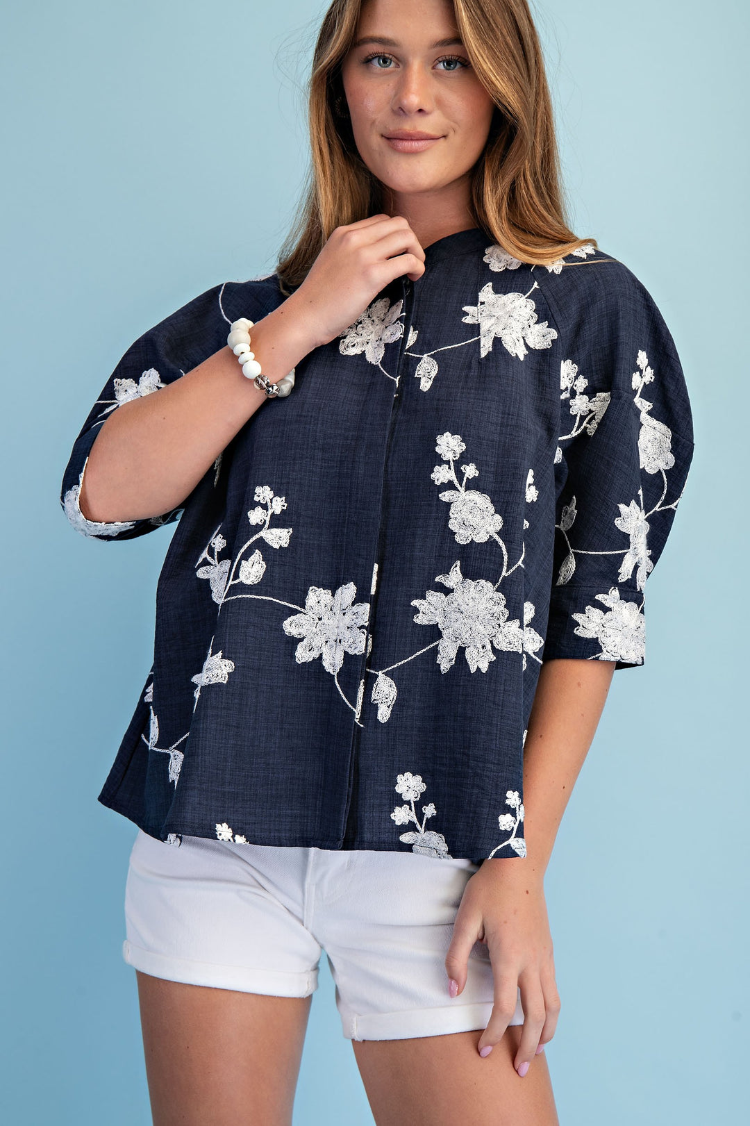 Woman wearing a navy blue floral blouse with white shorts against a light blue background