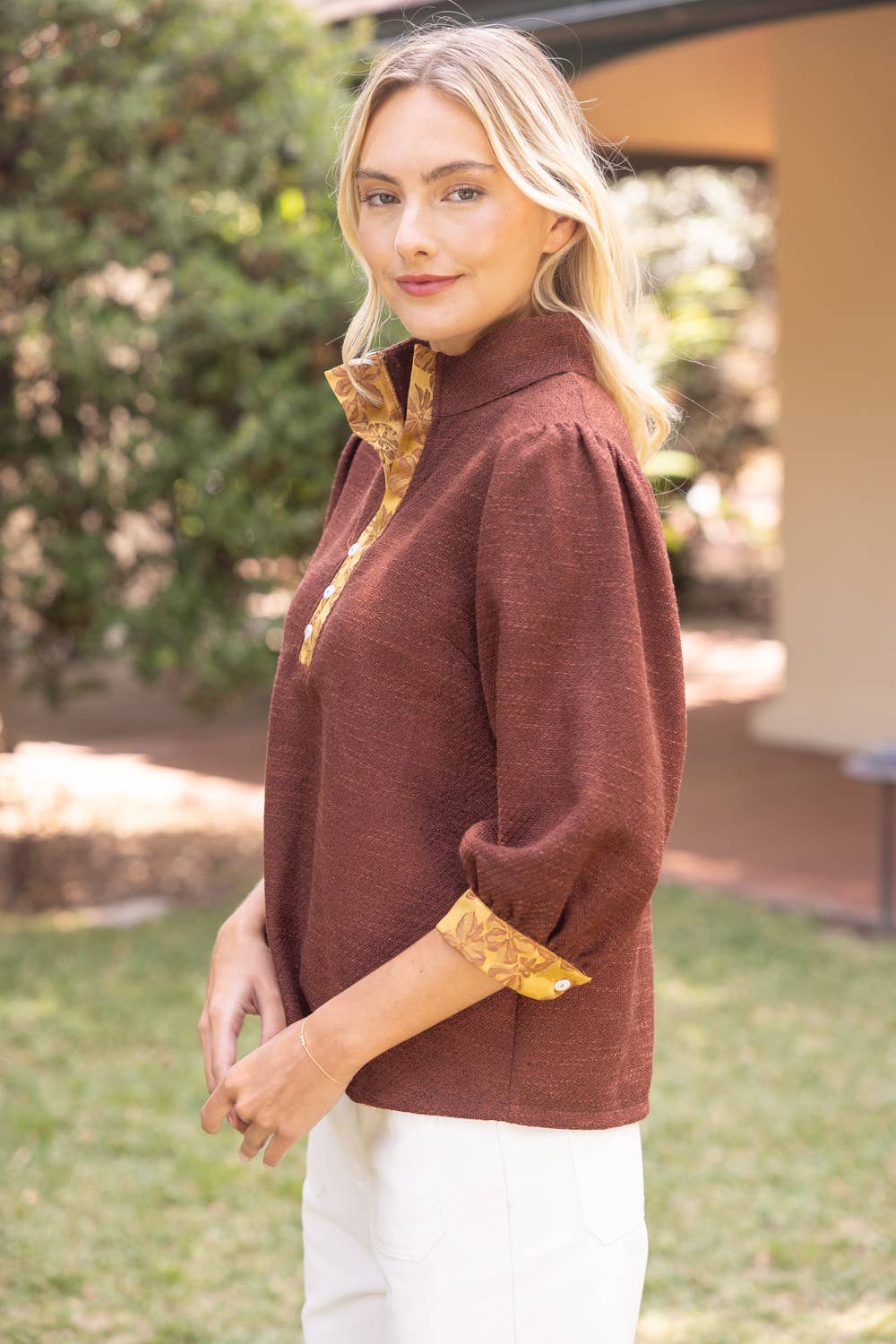 A woman wearing a rust-brown textured top with gold patterned trim on the collar and cuffs, paired with white pants, standing outdoors in front of a house and greenery.