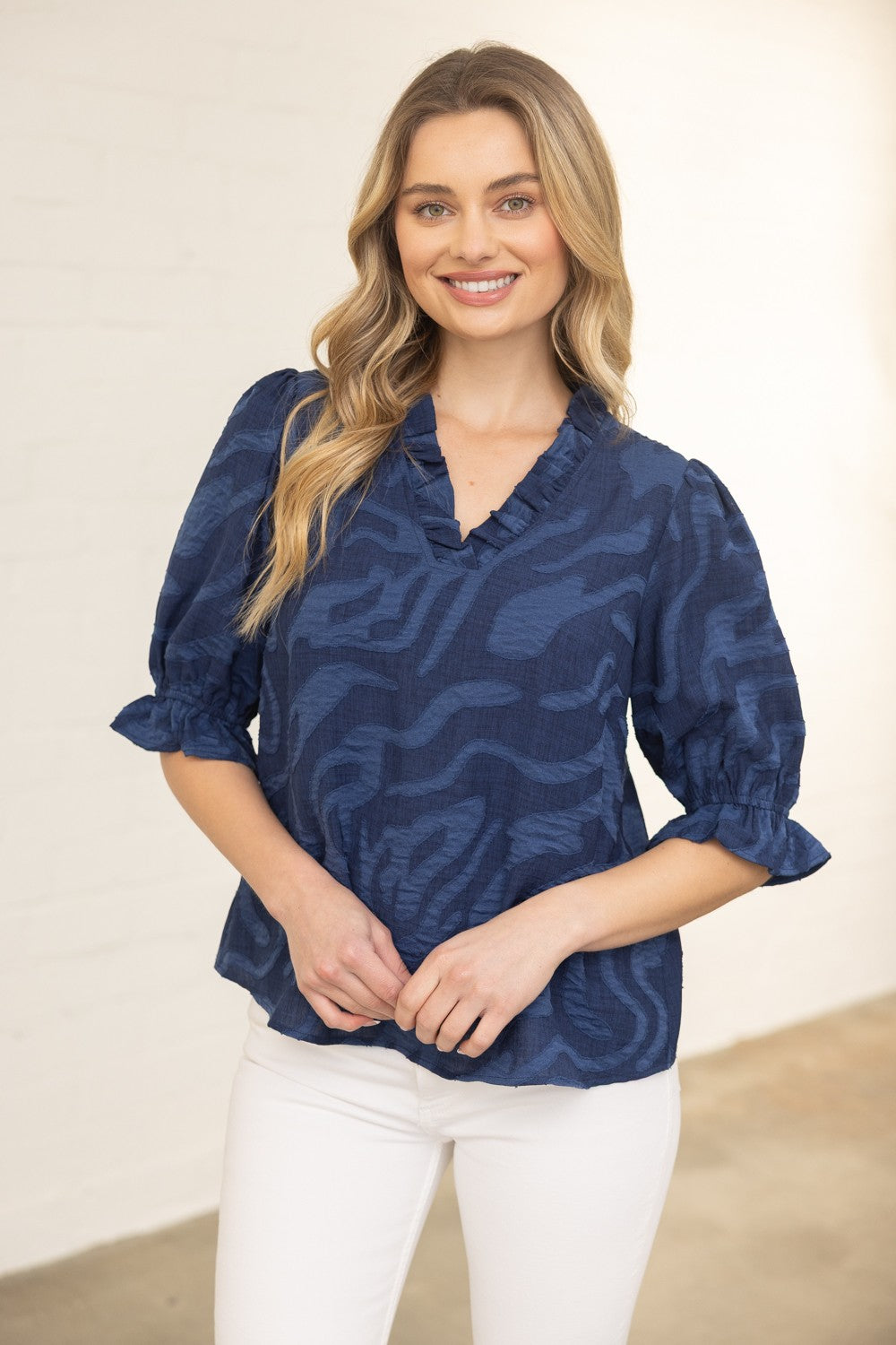 Woman wearing a blue patterned blouse and white pants against a beige background