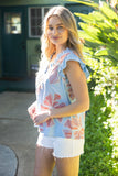 Woman wearing a floral top and white shorts standing outdoors with greenery in the background