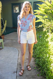 Woman in a floral top and white shorts standing outdoors near a green door.