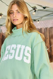Person wearing a light green sweatshirt with 'JESUS' printed on it, standing indoors.