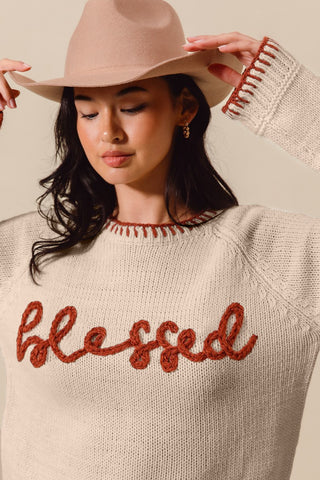 Woman wearing a beige sweater with 'blessed' text and a matching hat against a neutral background