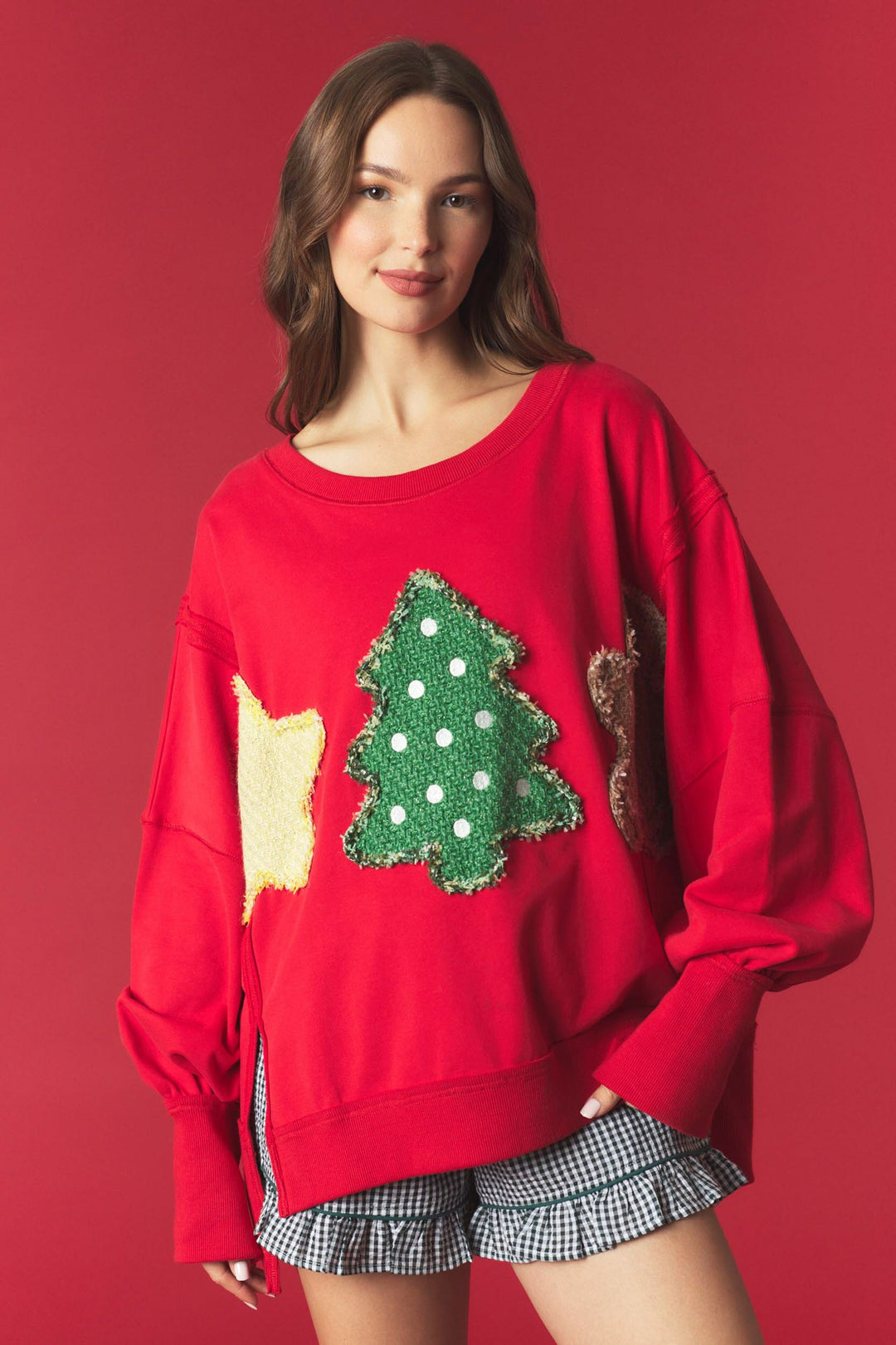 A woman modeling a festive red crewneck sweatshirt with a green sequined Christmas tree and a yellow sequin star on the front, paired with gingham ruffled shorts, set against a solid red studio background.
