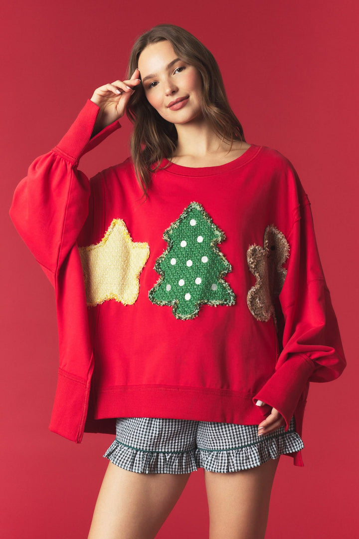 A woman modeling a festive red crewneck sweater with applique patches of a yellow star, green Christmas tree, and brown squirrel, paired with blue and white gingham ruffled shorts, set against a solid red studio background.